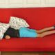 Den Kopf mit Kissen verdeckt liegender Junge auf rotem Sofa